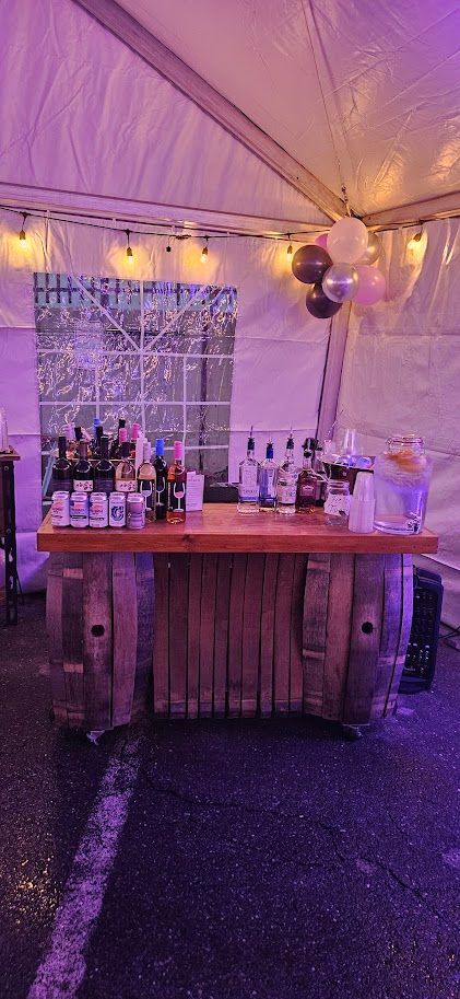 Decorated outdoor tent with a wooden bar table, bottles, and purple lighting for an event