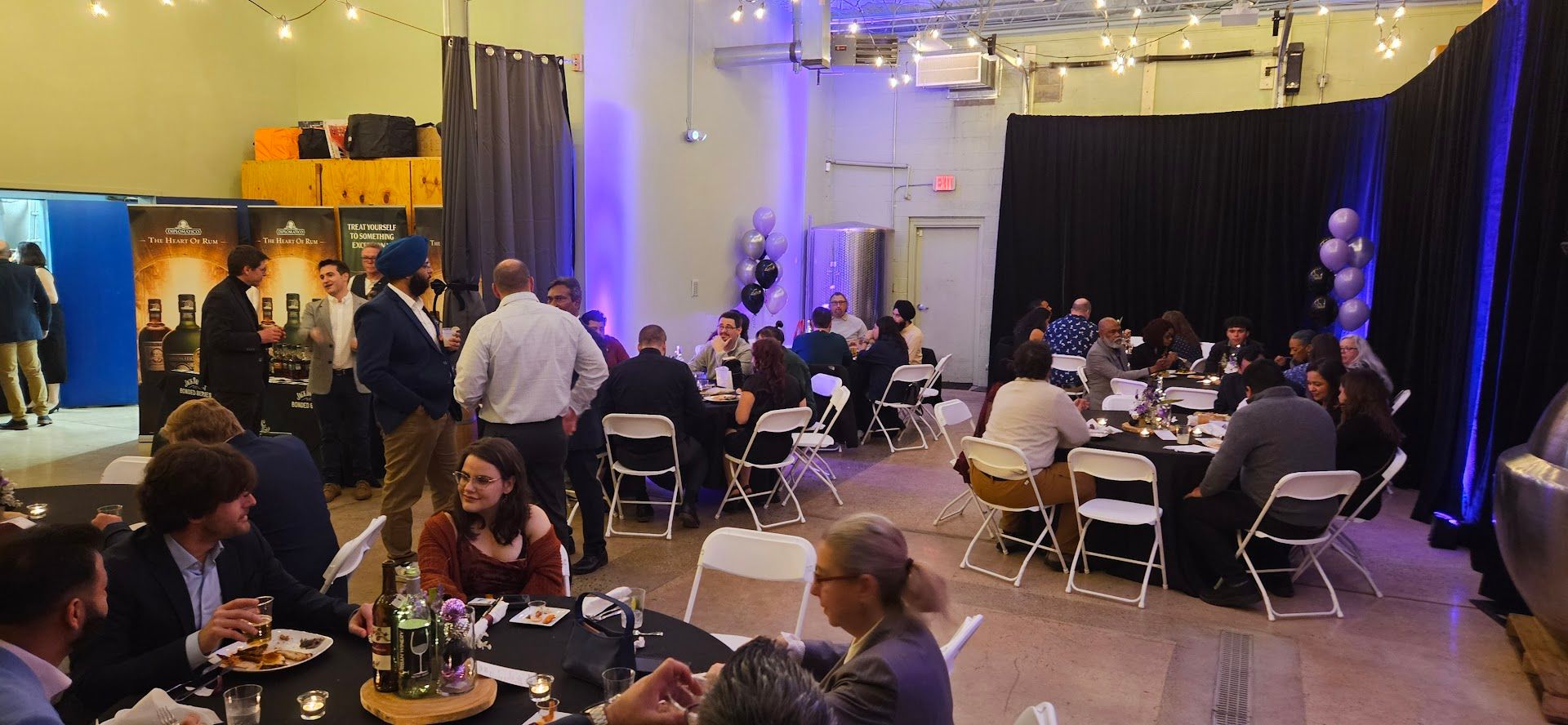 Guests seated at round tables in a banquet hall, with a lit stage and formal event lighting in the background