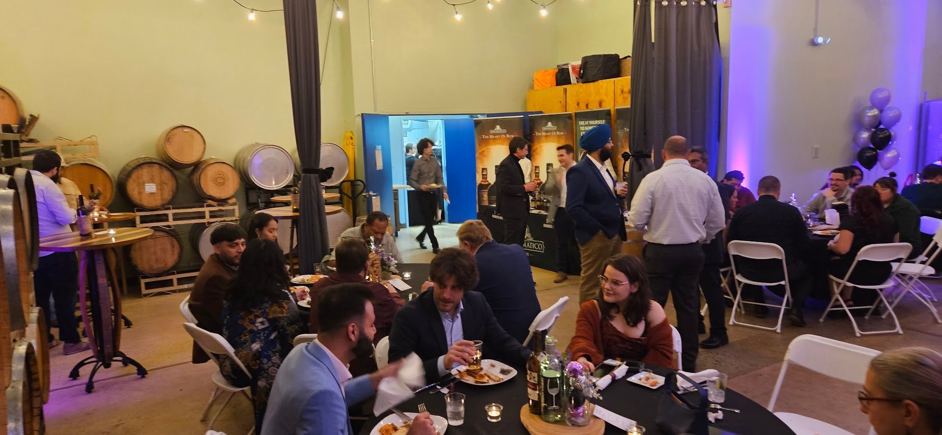 People mingling at a lively indoor event with tables, drinks, and barrels in the background