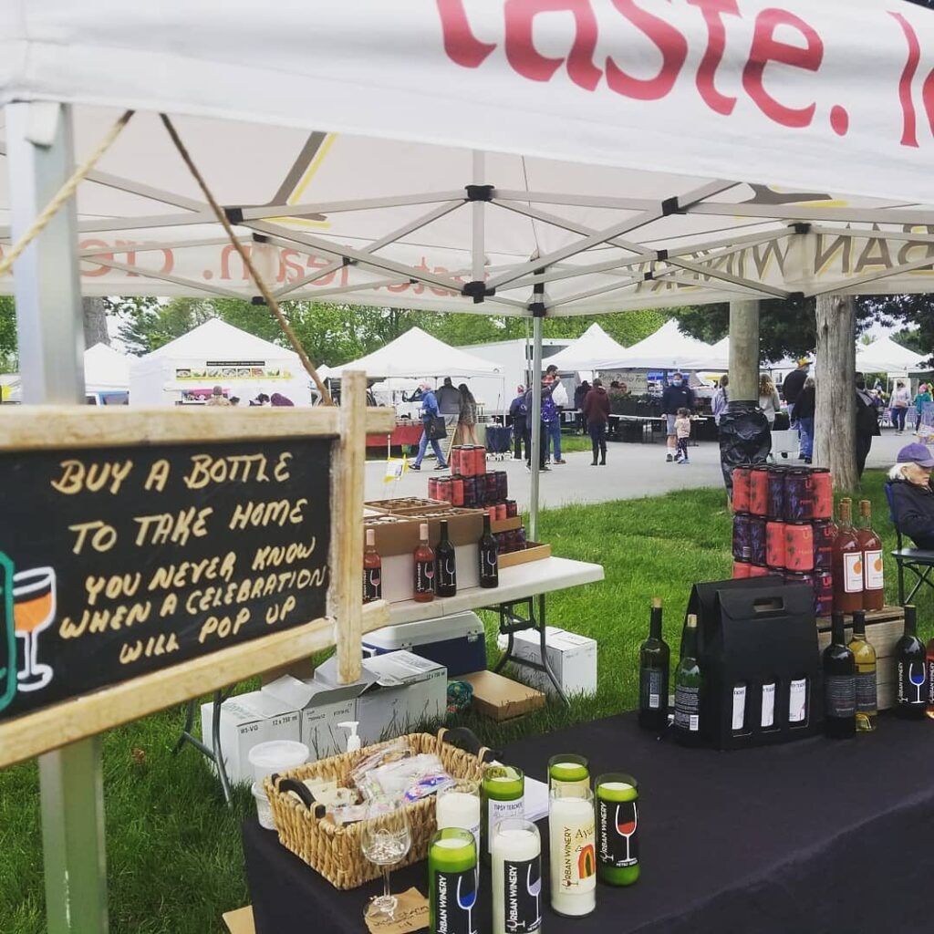 Outdoor market booth under a white canopy with bottles, jars, and a chalkboard sign about buying a bottle to the home team.