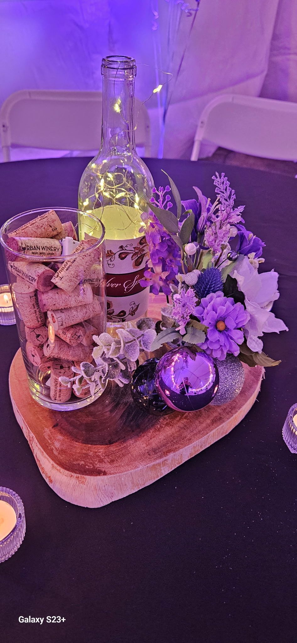 Flower arrangement and glass bottle centerpiece on a wooden slab, lit with purple ambient light.