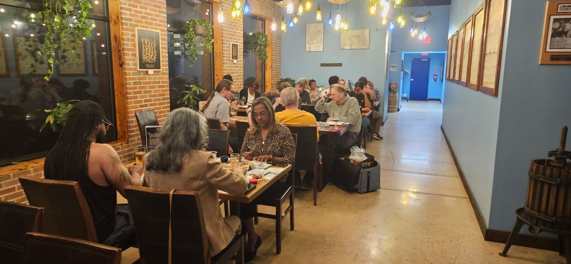 Cozy indoor restaurant with diners seated at wooden tables under warm hanging lights and greenery.