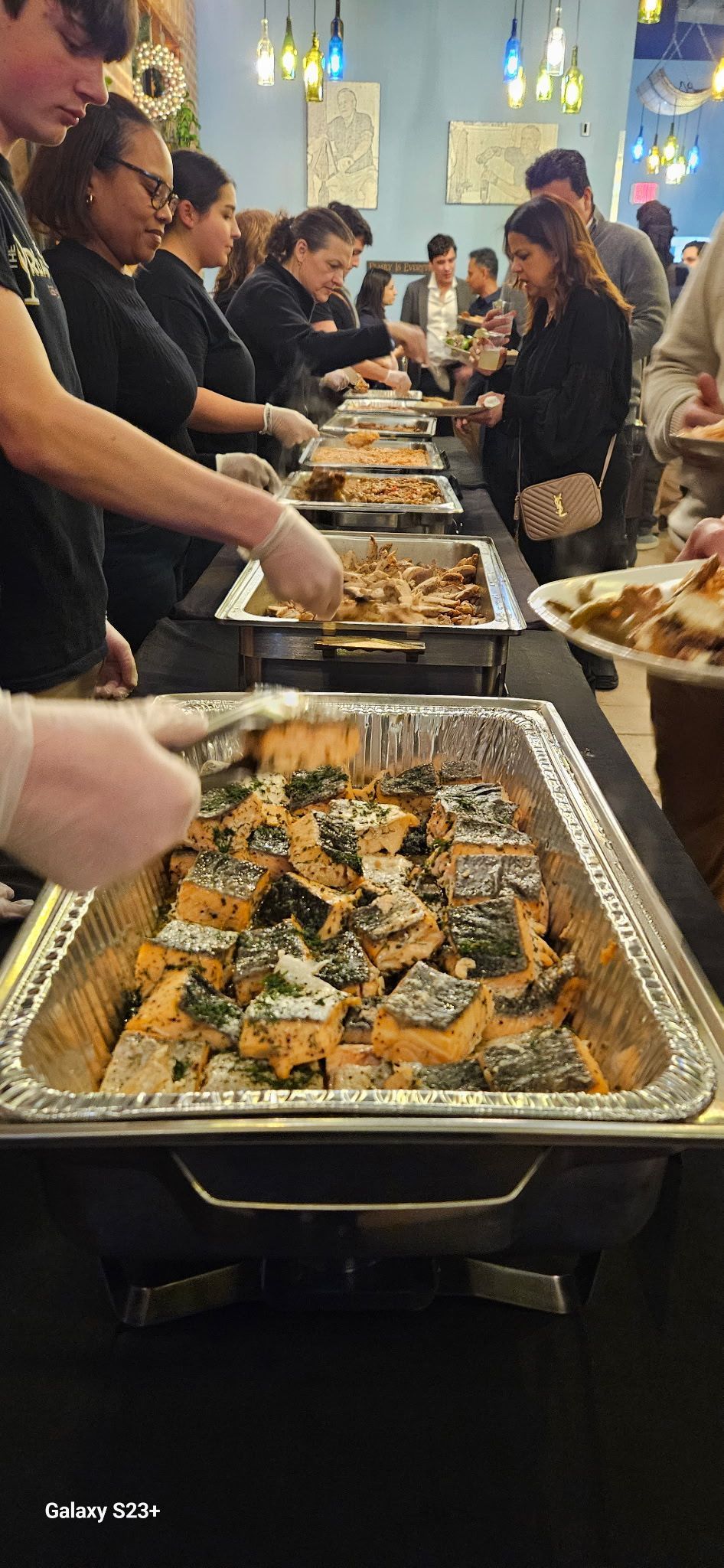 Buffet line with people serving themselves from trays of baked food in a warmly lit indoor hall
