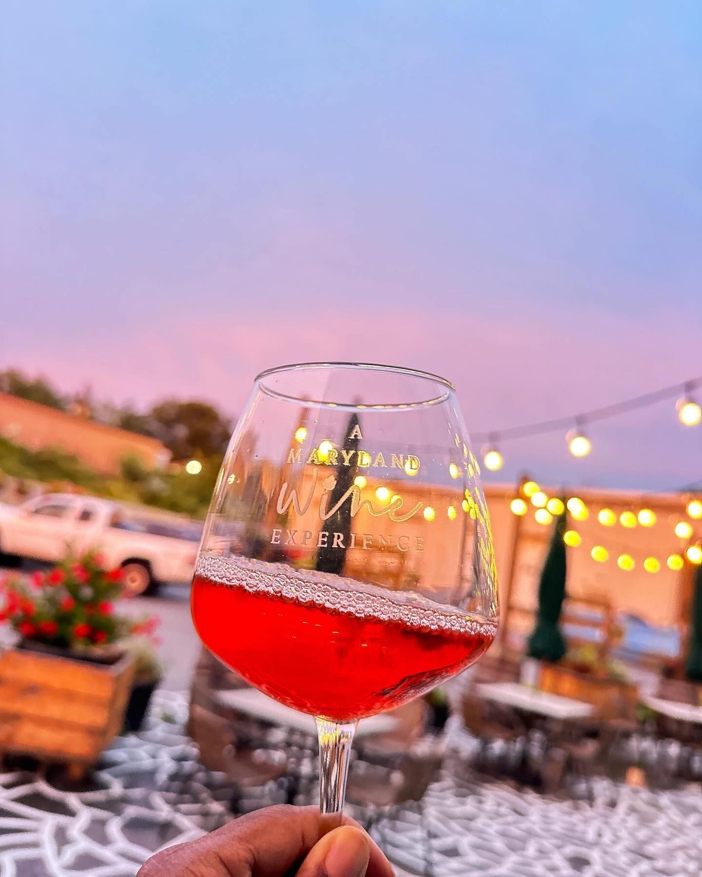 Wine glass with red drink held up at an outdoor evening patio with string lights and a pastel sunset sky