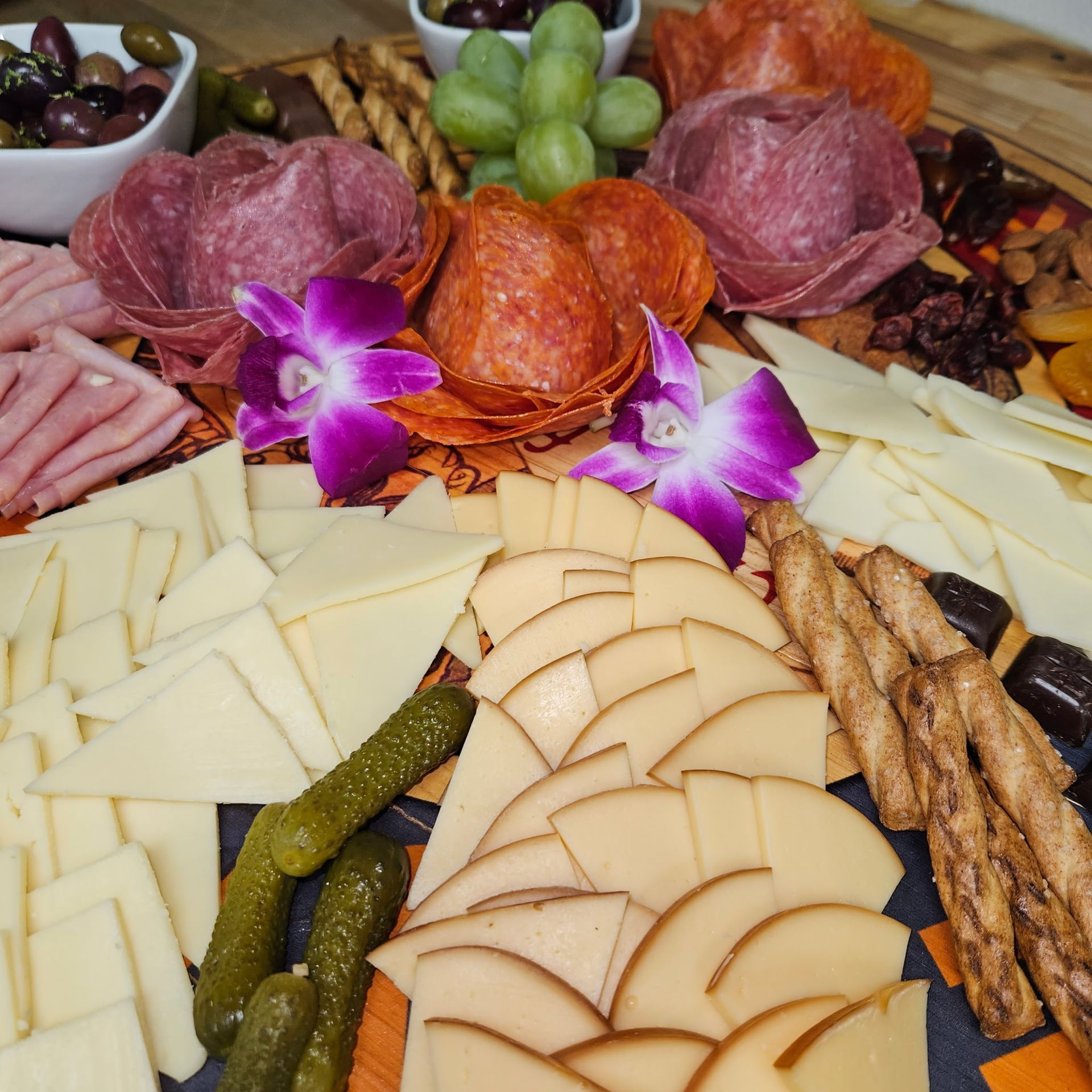 Charcuterie board with sliced meats, cheeses, grapes, pickles, and breadsticks, garnished with purple orchids