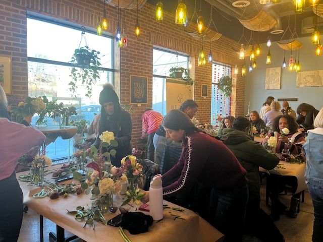 People arranging flowers at long tables in a bright workshop with hanging lights and large windows