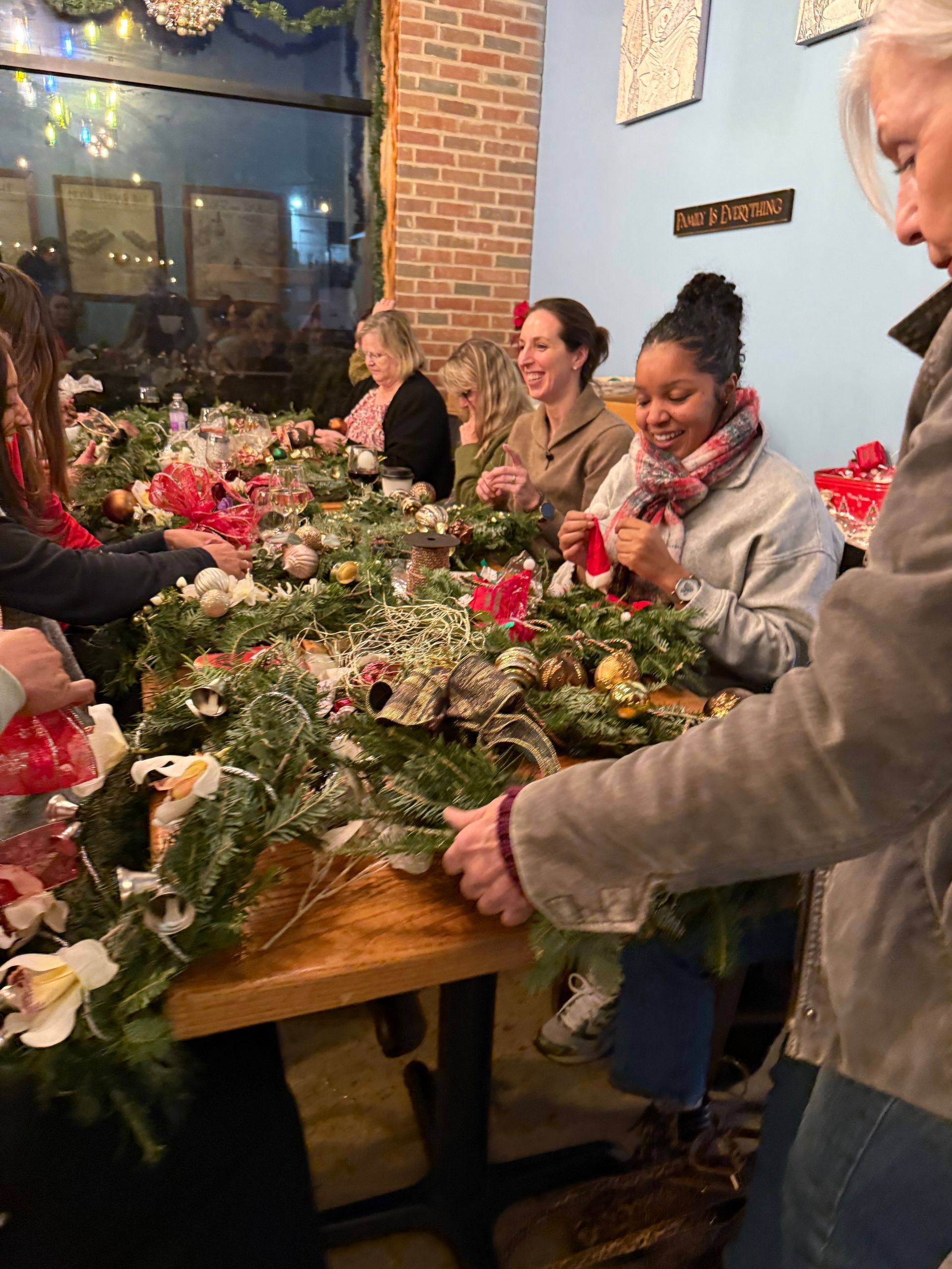 People arranging festive holiday wreaths around a wooden table in a warmly lit room