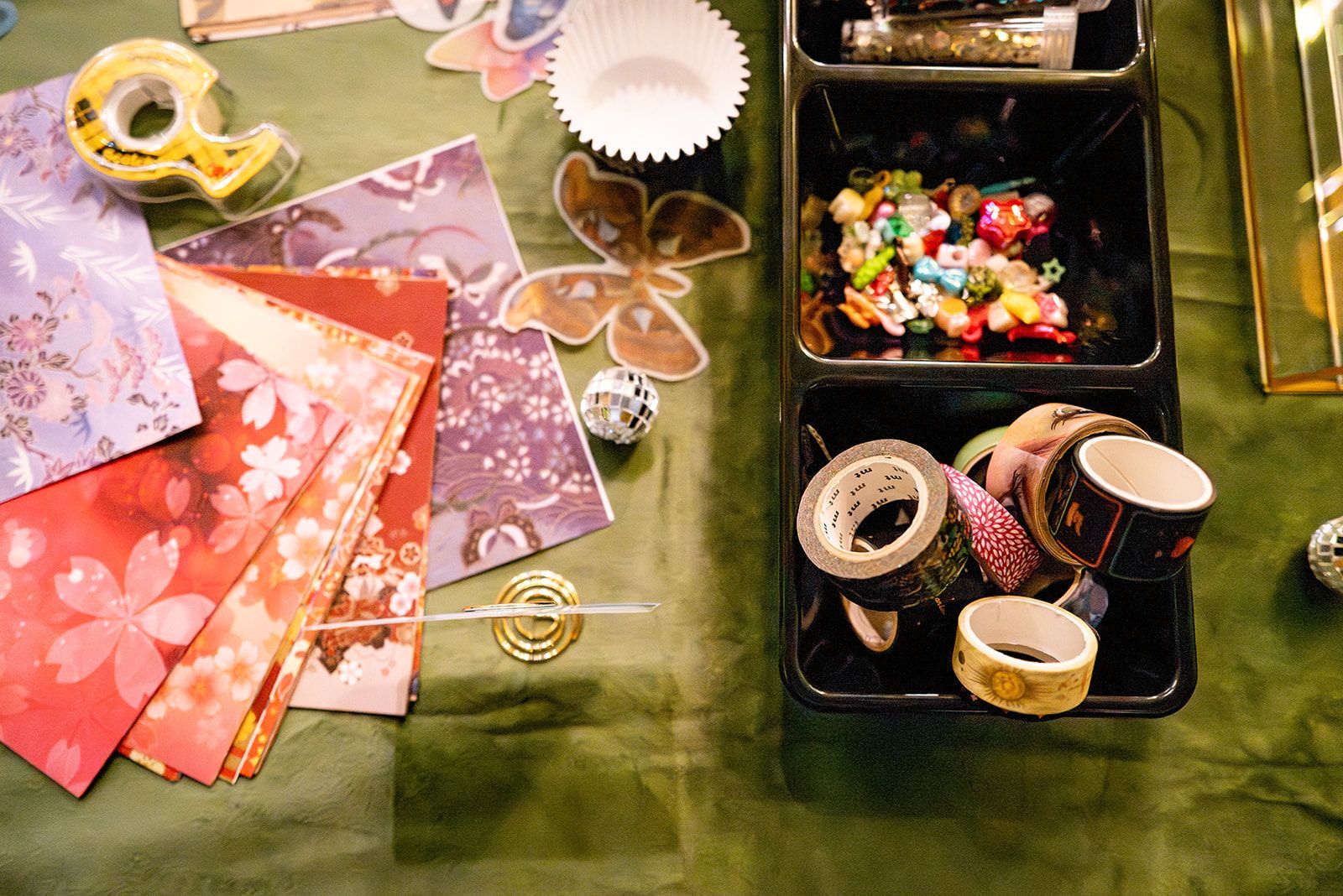 Craft supplies on a green table: patterned paper, tape, buttons, ribbon, and small containers of embellishments.