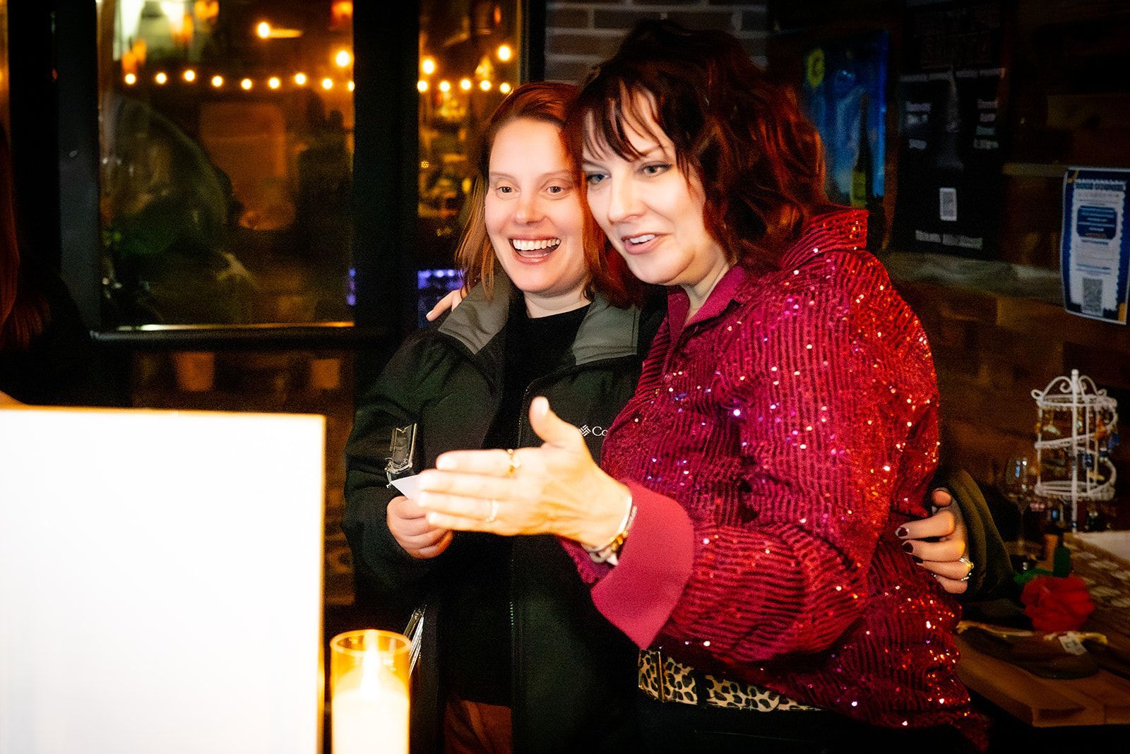 Two people smiling together in a cozy, dimly lit bar, one in a red sequined top gesturing toward the camera