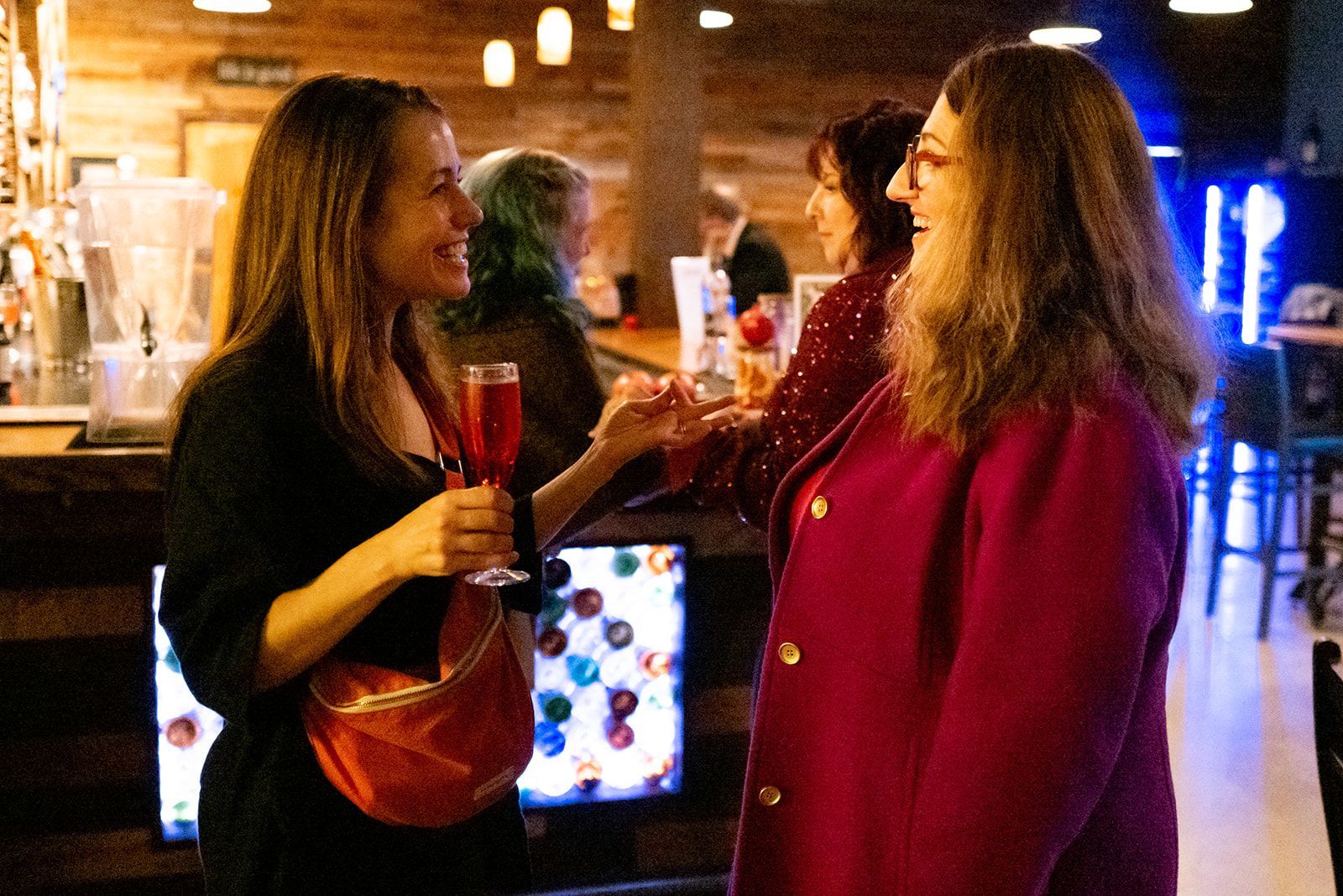 Two people chatting and smiling at a dimly lit bar, holding drinks near a glowing display.