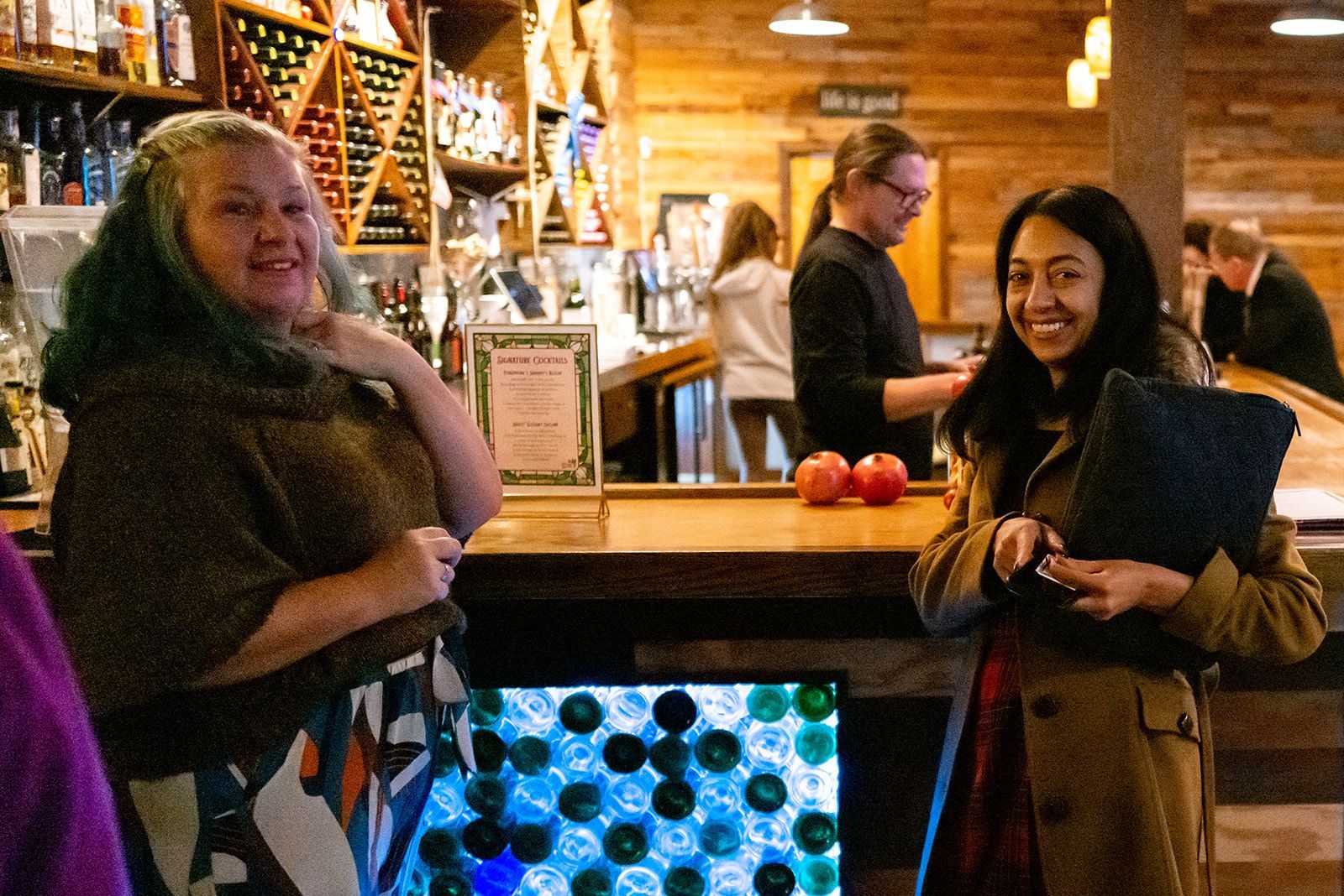Two people standing at a bar with warm lights and bottles in the background