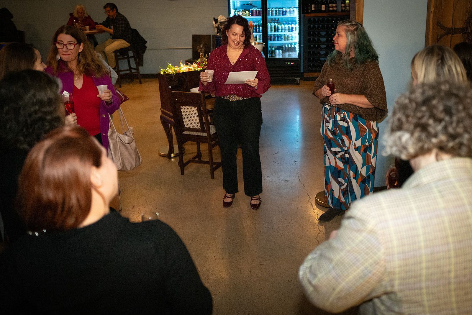 People gathered in a room, with three women standing and speaking to a small seated audience.