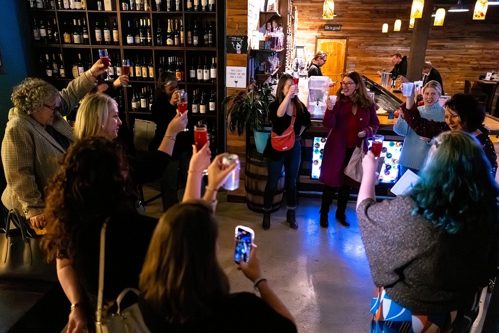 People gathered in a dim bar or shop, raising glasses and phones during a small celebration.