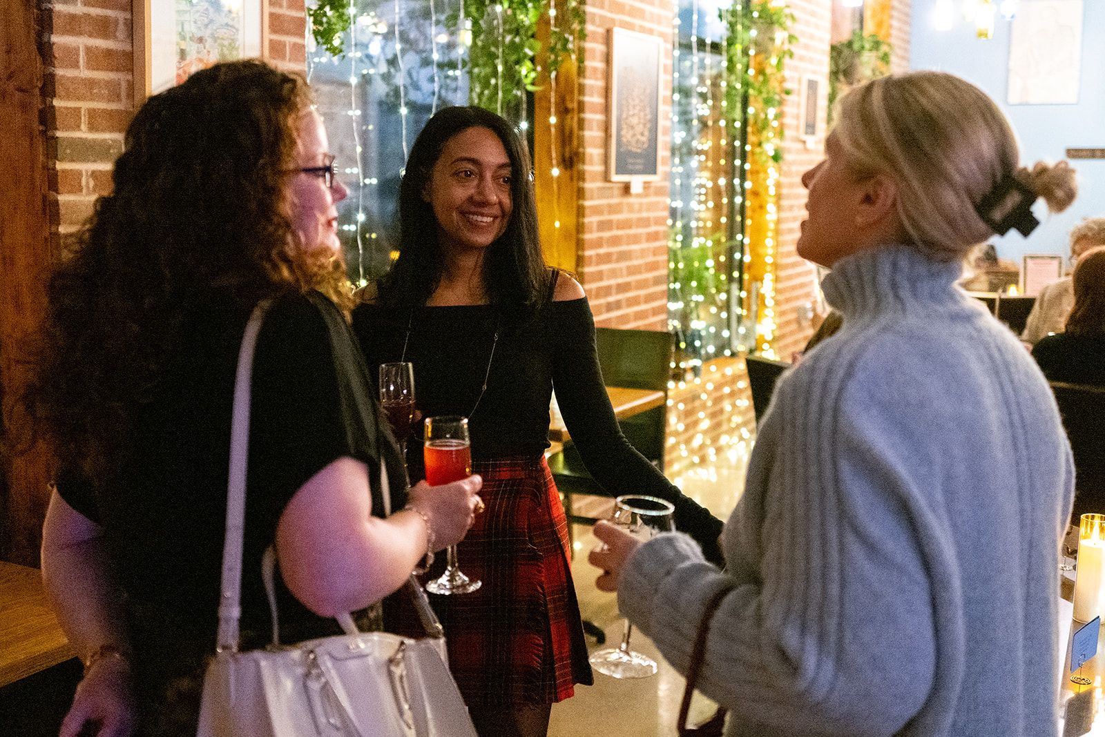 Three people chatting outdoors at a warmly lit party, holding drinks near a brick building.
