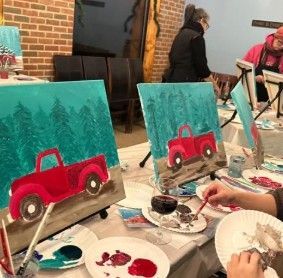 People painting red trucks on teal canvases at an art class table
