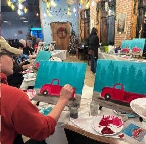 People painting red pickup trucks on blue canvases at a festive art class table