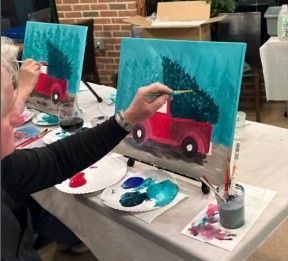 People painting red trucks on teal canvases at an art studio table with paint palettes