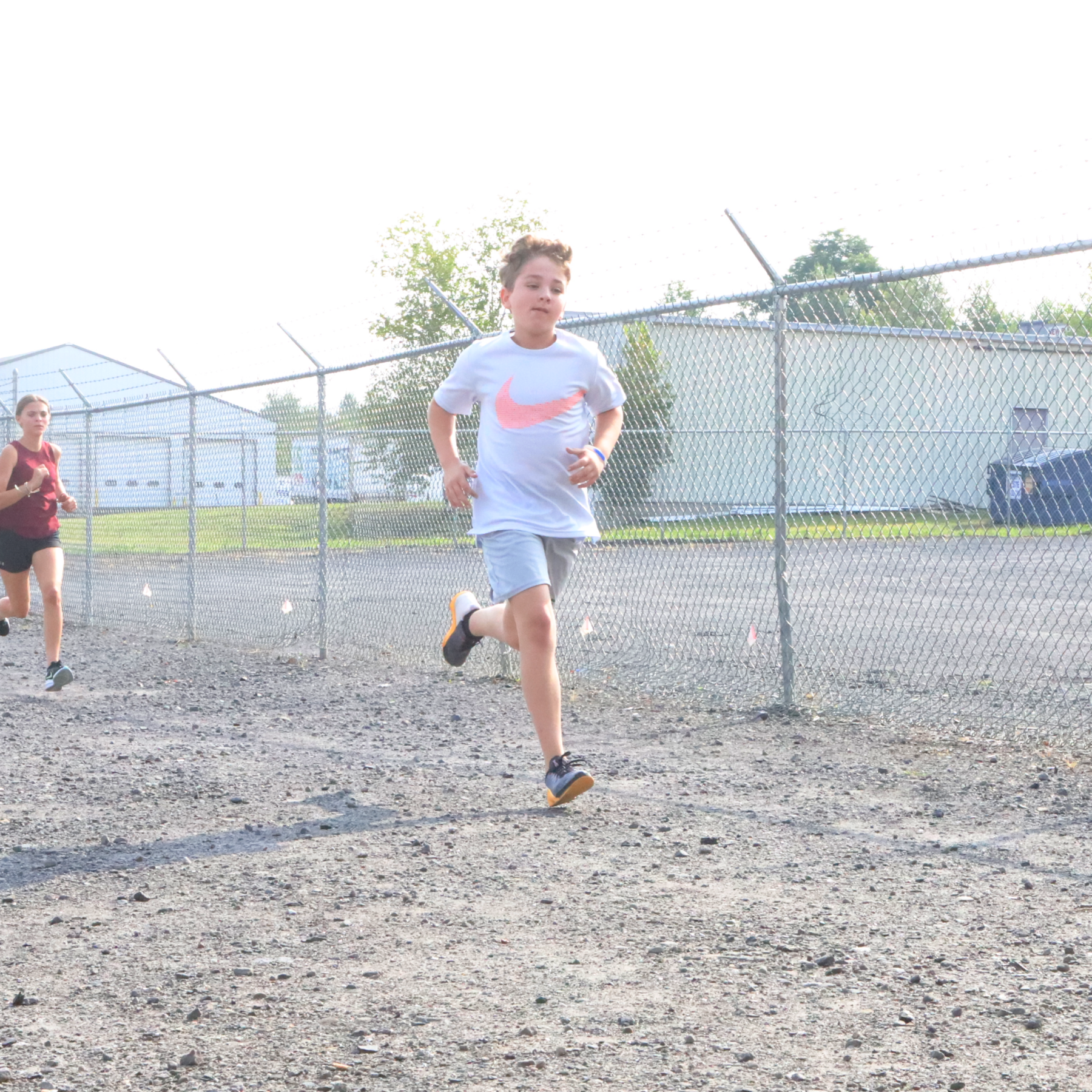 A boy and a girl are running in a dirt field behind a chain link fence.