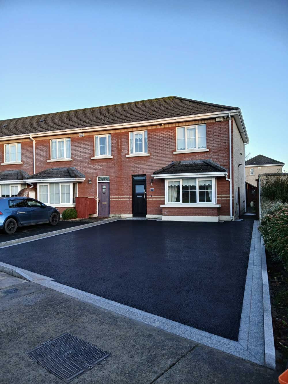 Black paved surface in front of a brick building with windows. Bordered by brick and greenery.