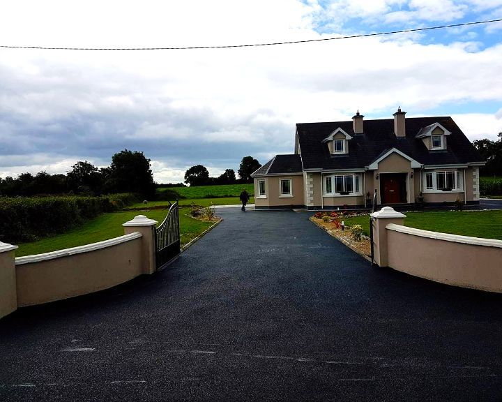 Black asphalt driveway leading to a light beige house with a dark roof. Person walks towards the house. Green fields.