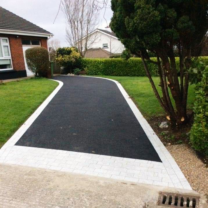 Black asphalt driveway with white brick border, green lawn, and trees.