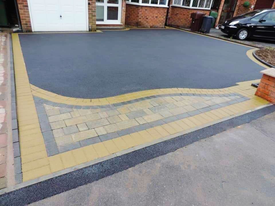 Driveway paved with dark asphalt, bordered by yellow and gray paving stones. Garage door in background.