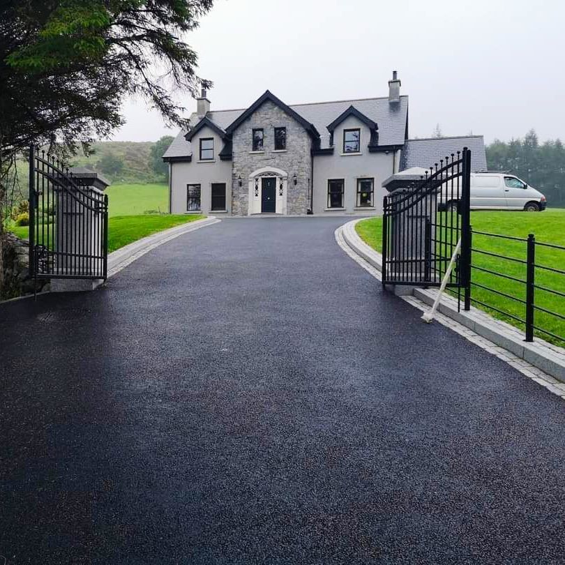 A gray stone house with a black asphalt driveway, black gates, and green lawn.