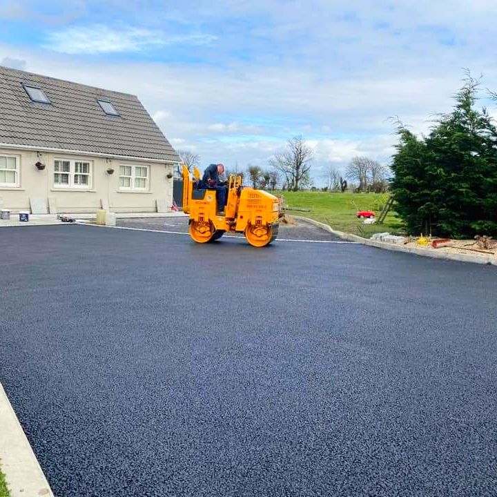 Asphalt driveway being rolled by a small yellow machine near a house with a green lawn.