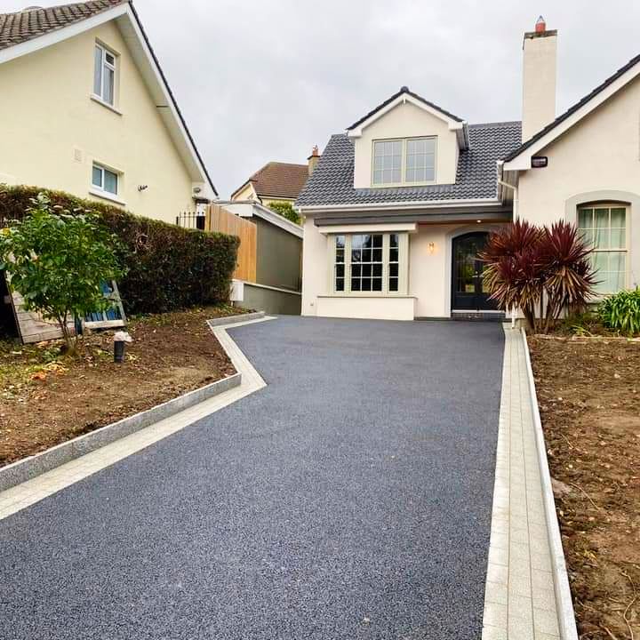 Driveway of a home, dark gray asphalt, with light-colored edging. Two-story beige house in background.