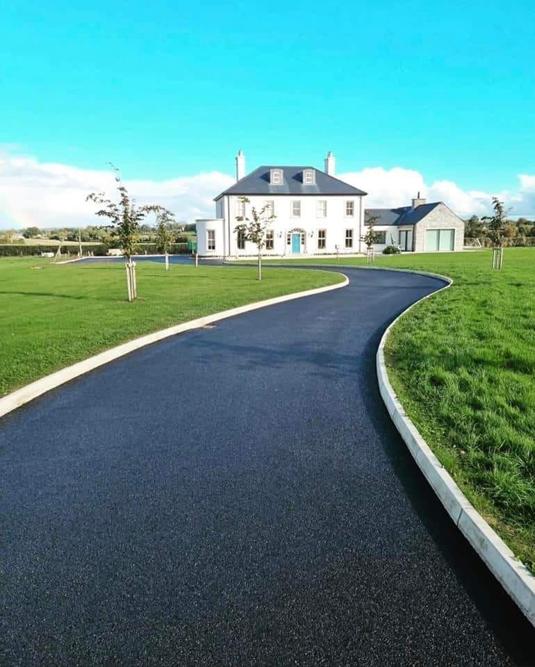 A long, black asphalt driveway curves towards a white house with a blue door, set on green grass under a blue sky.