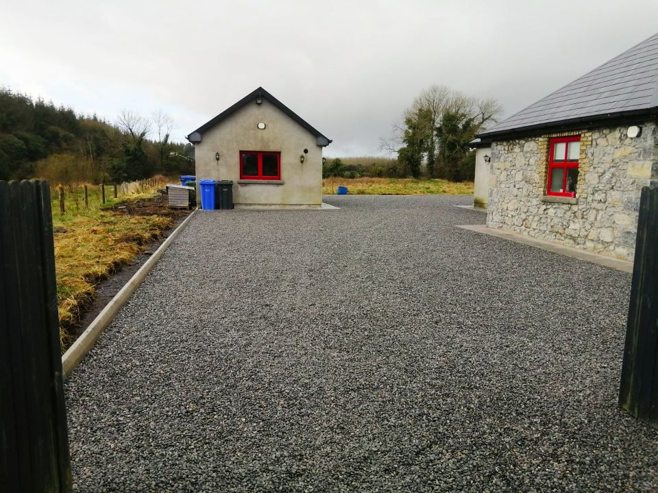 Gravel driveway leads to two buildings with red window frames. Overcast sky, field and fence.