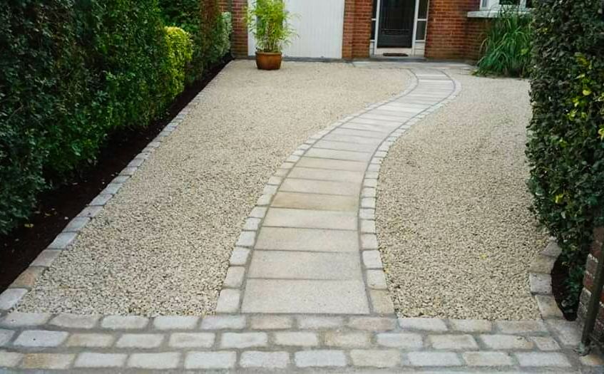 Stone pathway curves through gravel driveway, flanked by green hedges, leading to a house entrance.
