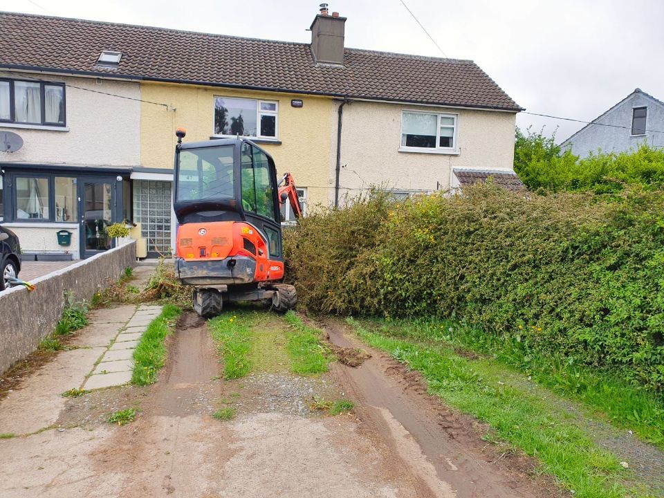 An orange excavator clearing a large hedge blocking a driveway in front of a house.
