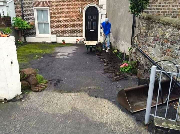 Man working on driveway with a wheelbarrow. Brick house, gray pavement, and debris are visible.