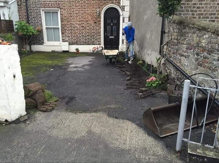 Man working on driveway with wheelbarrow and small excavator; brick building background.