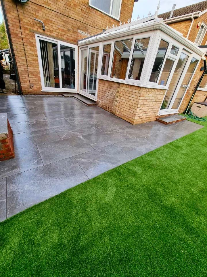 Gray patio with artificial green grass beside a brick house and white conservatory.