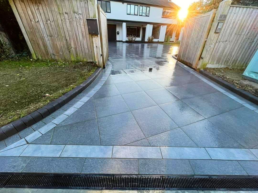 Driveway paved with grey blocks, bordered by a wooden fence, leading to a house, with sunlight.