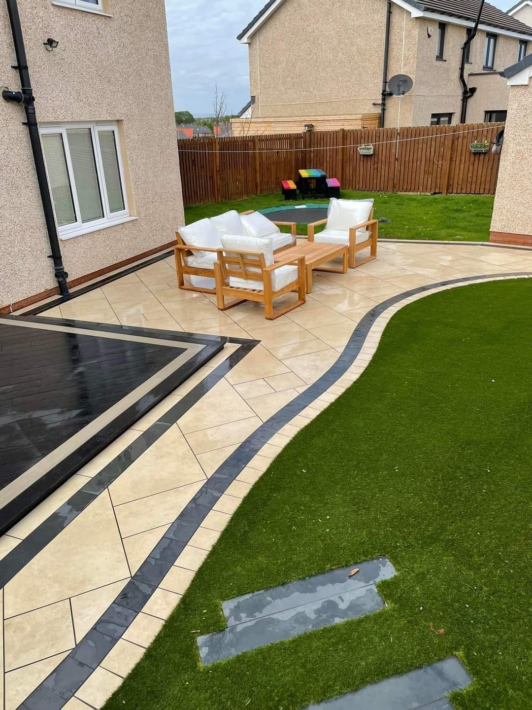 Patio with wooden furniture, bordered by black and beige paving, leading to a lawn.