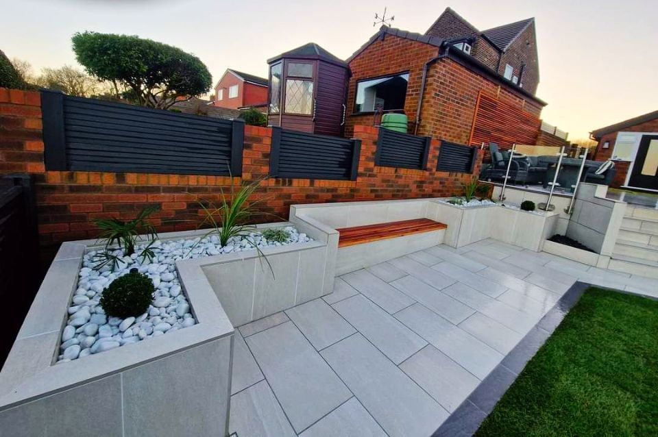 Modern patio with white pavers, built-in seating, and landscaped planters against a brick wall.