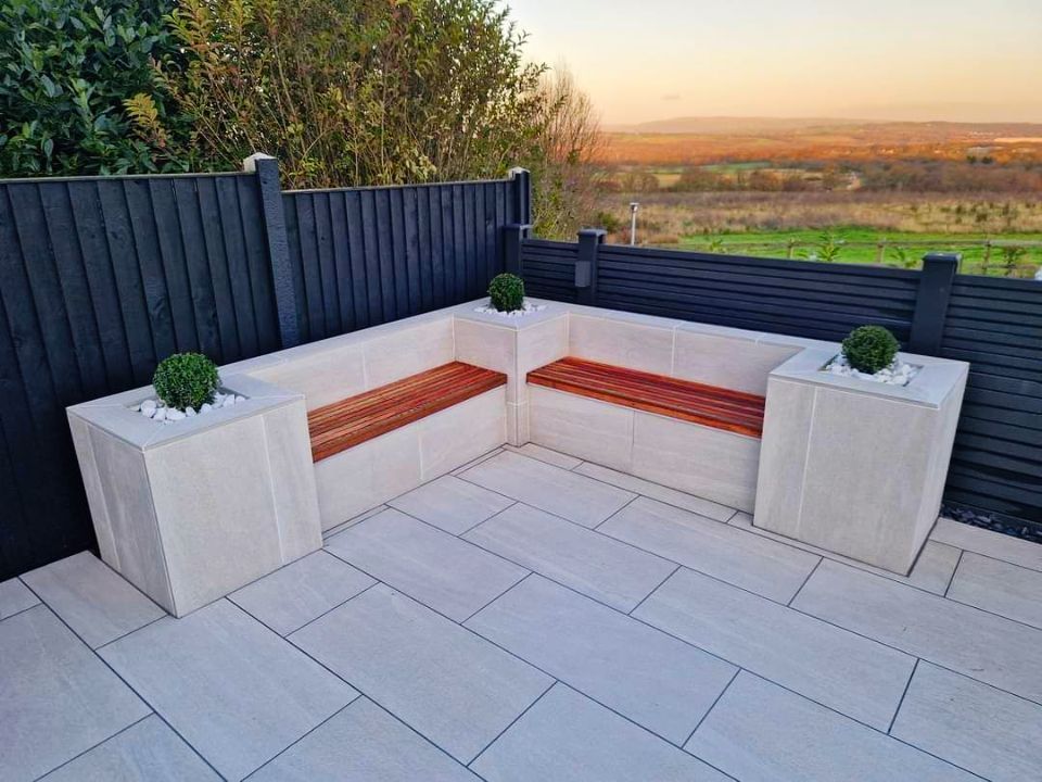 Corner outdoor seating with stone planters, wood bench, gray tile patio, black fence, and view of landscape.
