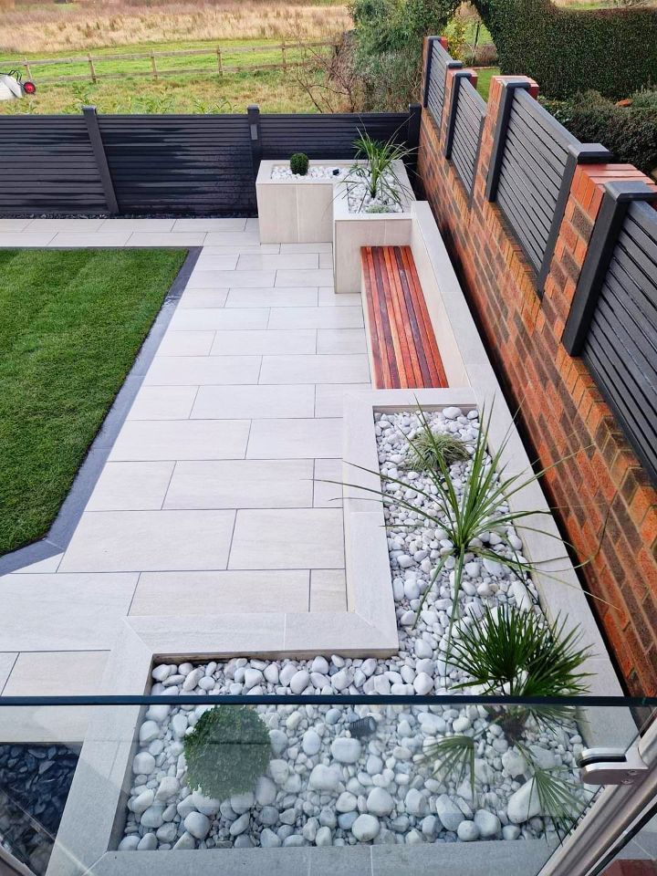Modern backyard patio with light stone pavers, a wooden bench, and white pebbles; a brick wall borders the right.