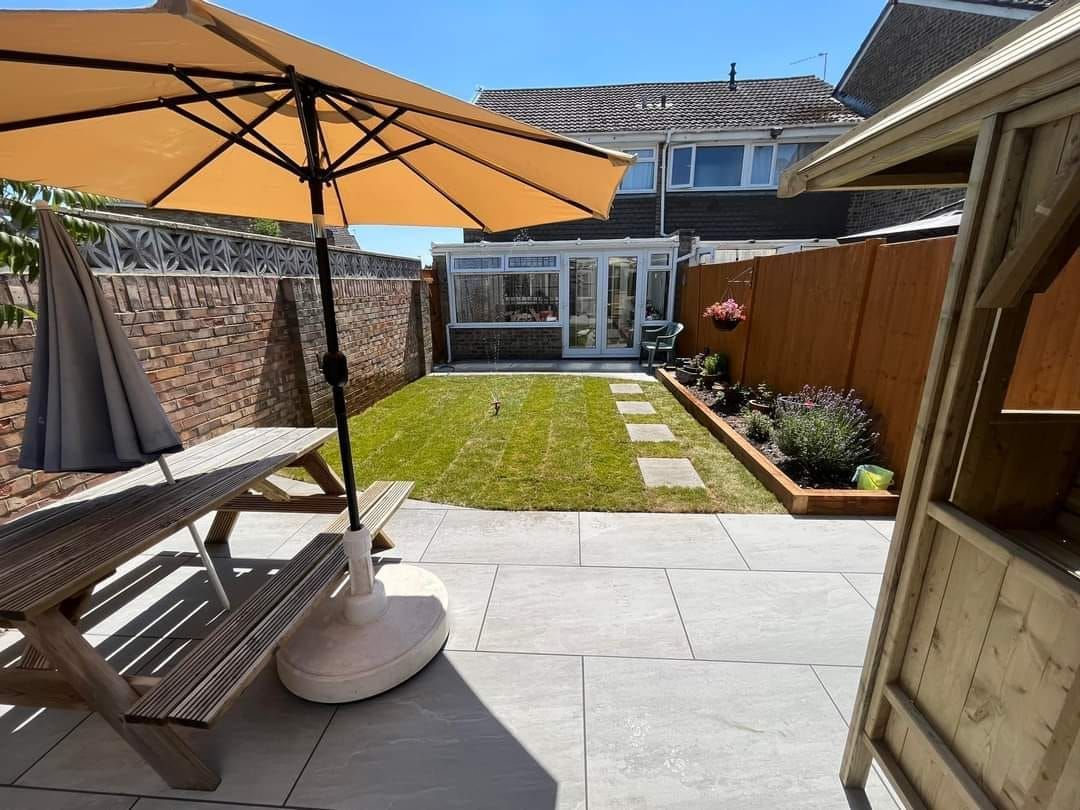 A backyard patio with a picnic table, umbrella, and a view of a lawn and a conservatory.