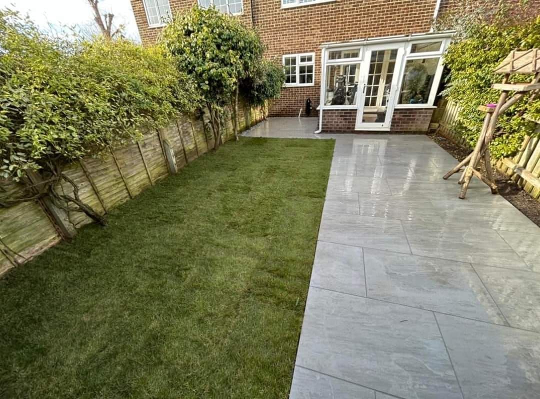 A backyard with a rectangular lawn and a gray tiled patio, next to a glass-walled sunroom and wooden fence.