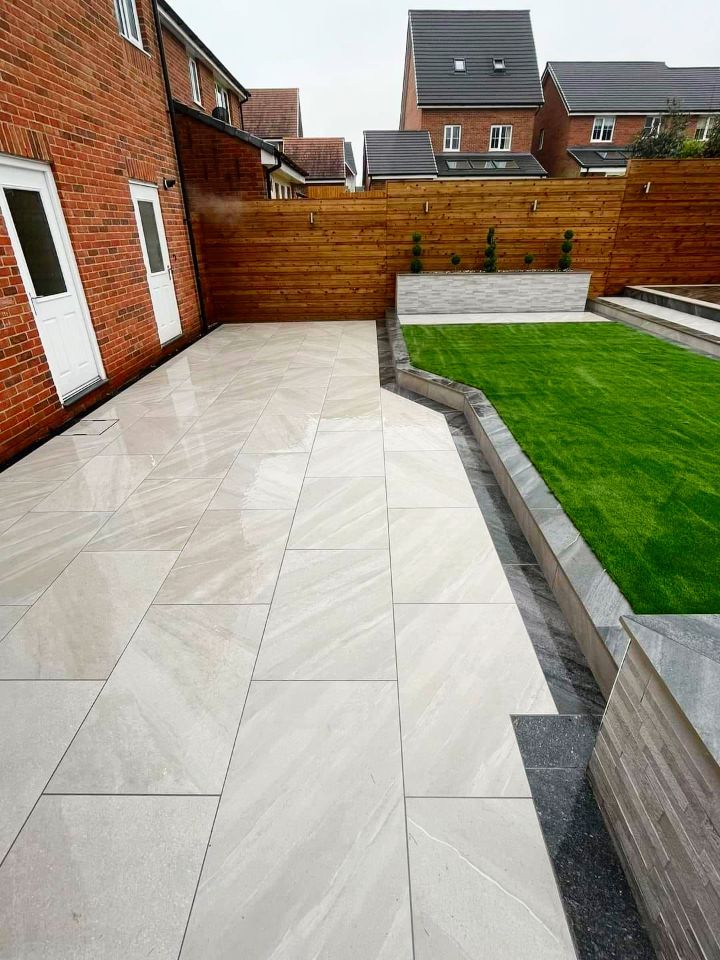 Patio with light stone tiles, green lawn, wooden fence, brick wall, and houses in the background.