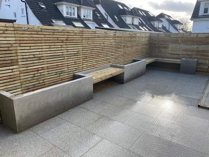 Patio with wood benches, metal planters, and a wooden slatted fence. Grey stone ground, cloudy sky.