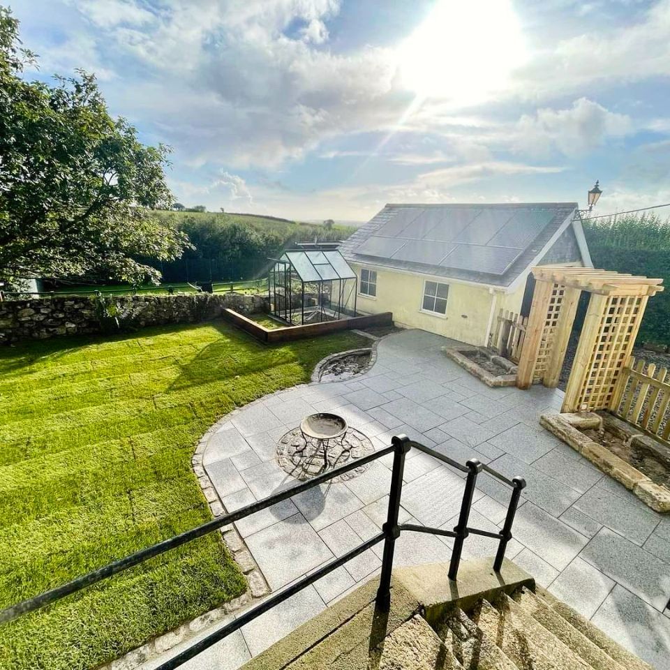 Backyard patio with fire pit, stone patio, green lawn, yellow house, and sunny sky.