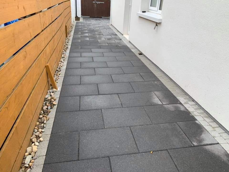 Dark rectangular paving stones form a walkway between a wooden fence and white building.