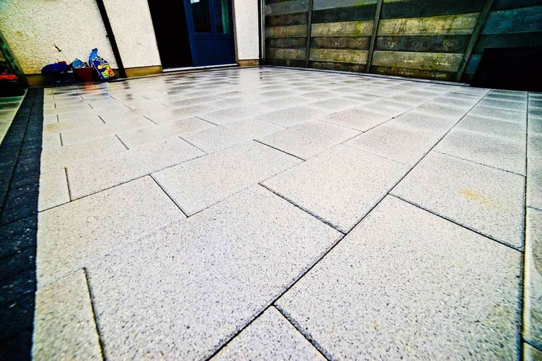 Gray stone patio with concrete block sections, near a building and fence.
