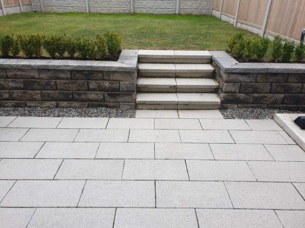 Stone patio with steps leading up to a raised garden bed with grass and shrubbery.