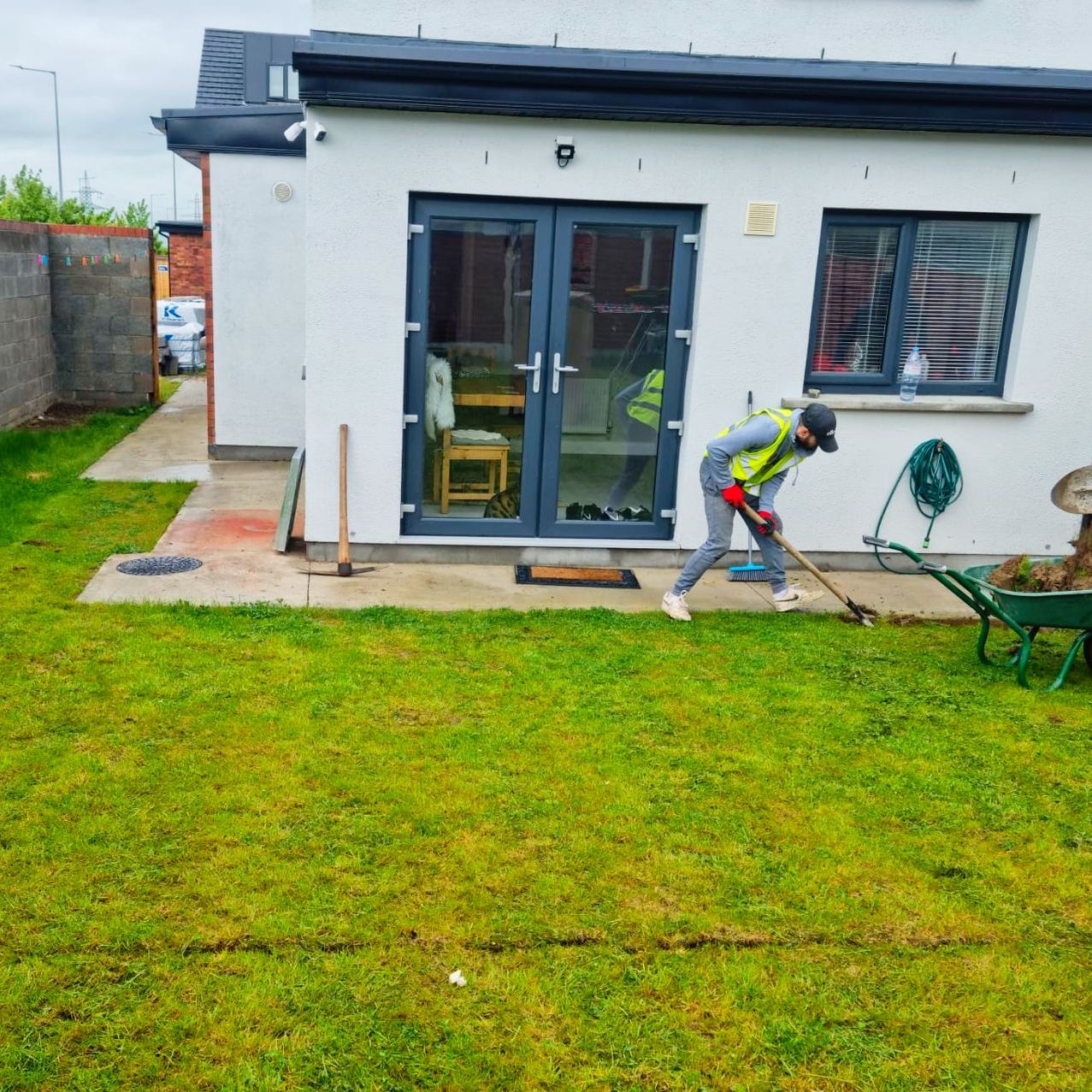 Person raking grass in a backyard. Building with glass doors, windows, and a wheelbarrow present.